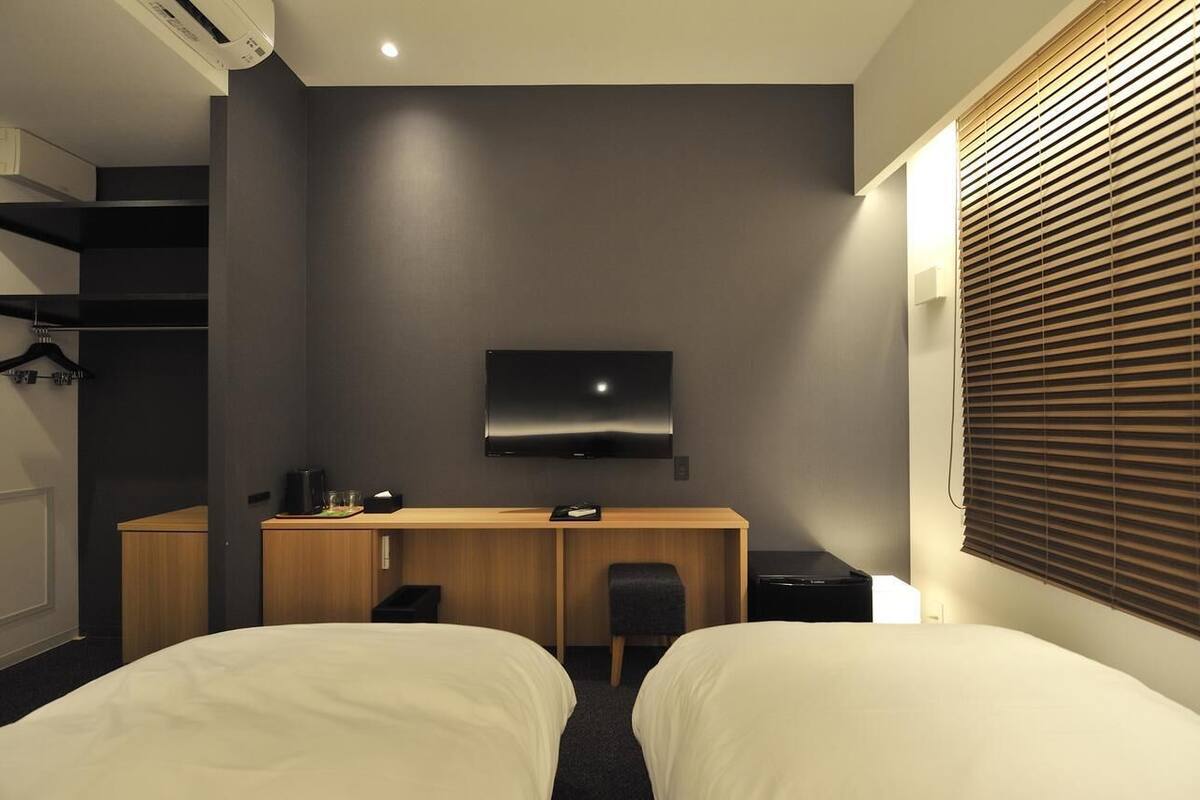A hotel room with a bed, a desk with a chair, a flat-screen TV mounted on the wall, and a window with blinds.