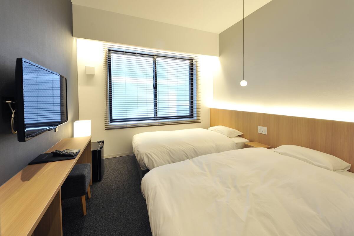 A hotel room with two beds, a desk, a TV, and a window with blinds.
