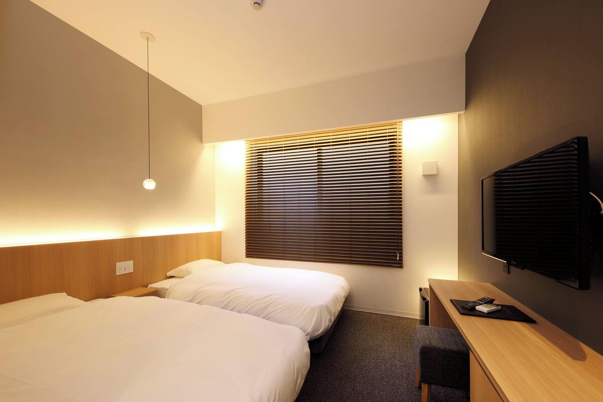 A hotel room with two beds, a desk, a television, and a window with blinds.