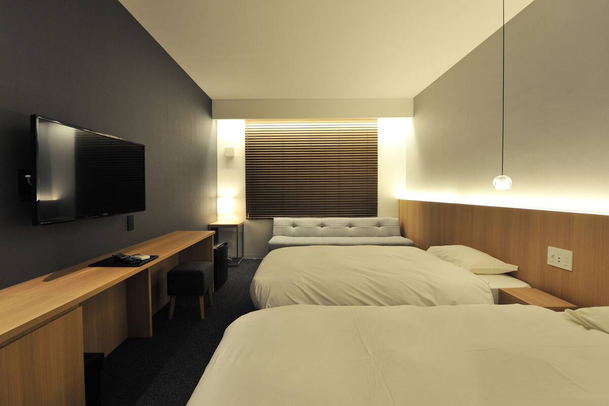 A hotel room with a bed, a desk, a TV, and a window with blinds.