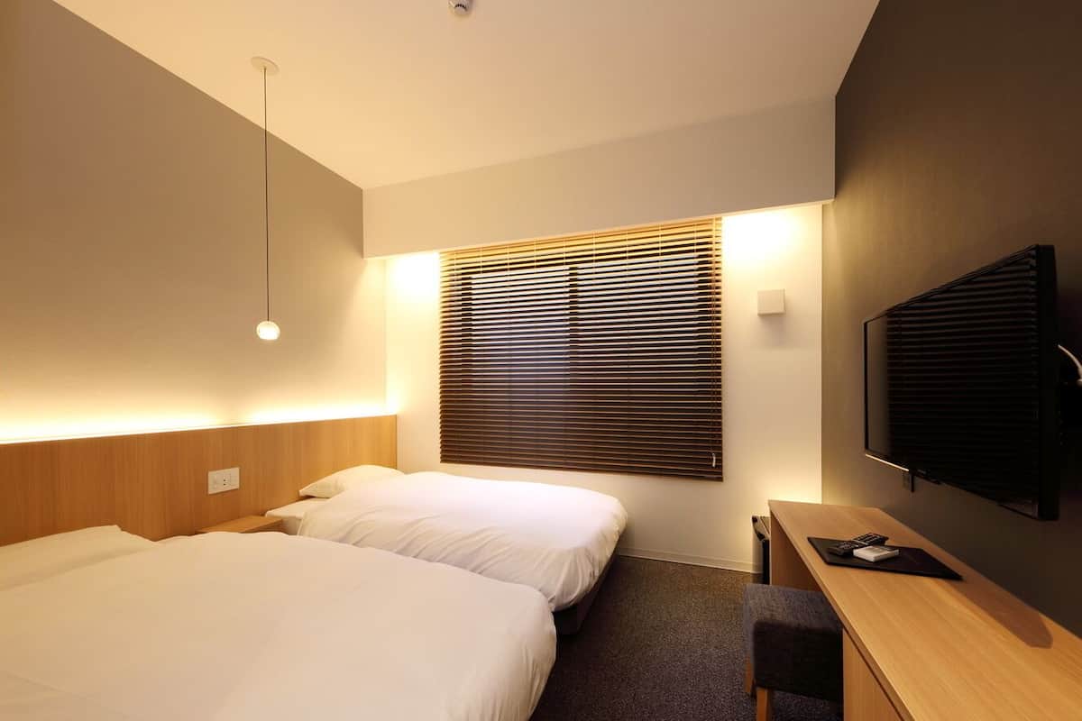 A hotel room with two beds, a desk, a television, and a window with blinds.