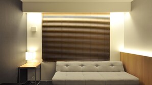 A hotel room with a bed, a sofa, a nightstand with a lamp, and a window with blinds. - FUGASTAY Shijo Omiya (Kyoto)