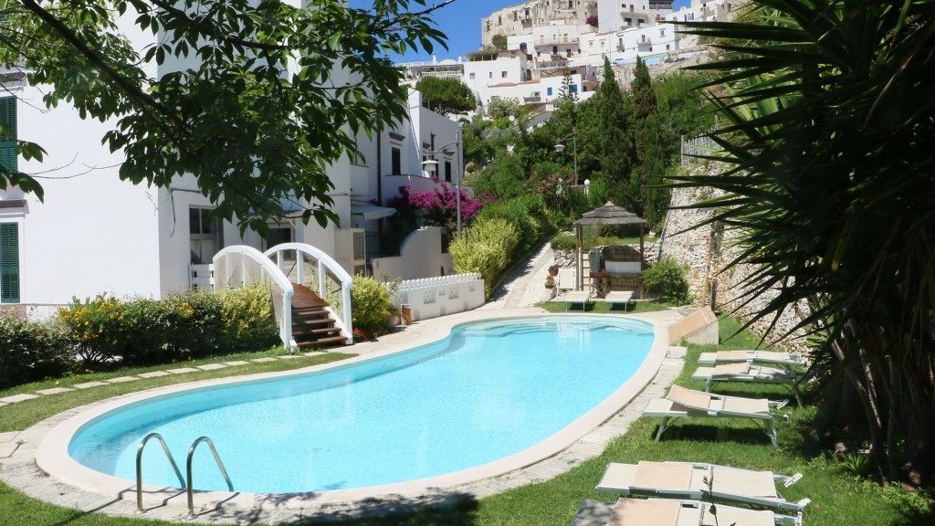 Una piscina de borde curvo, rodeada por un césped y un edificio blanco con balcones.