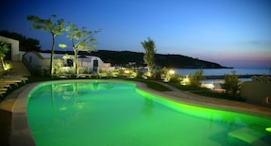 A swimming pool with a curved edge, surrounded by a landscaped area with trees and a view of a body of water in the background. - Hotel Elisa (Peschici)