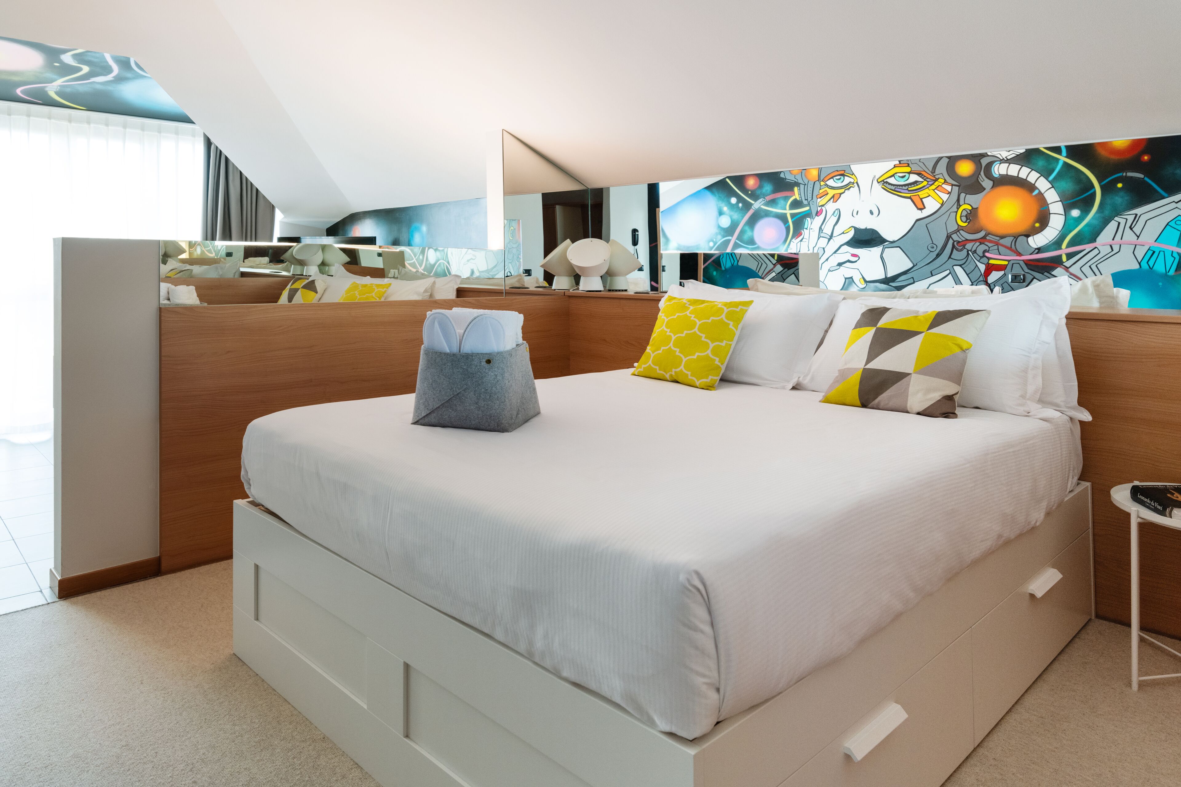 A modern hotel room with a large bed, a bedside table, a mirror, and a colorful abstract painting on the wall.