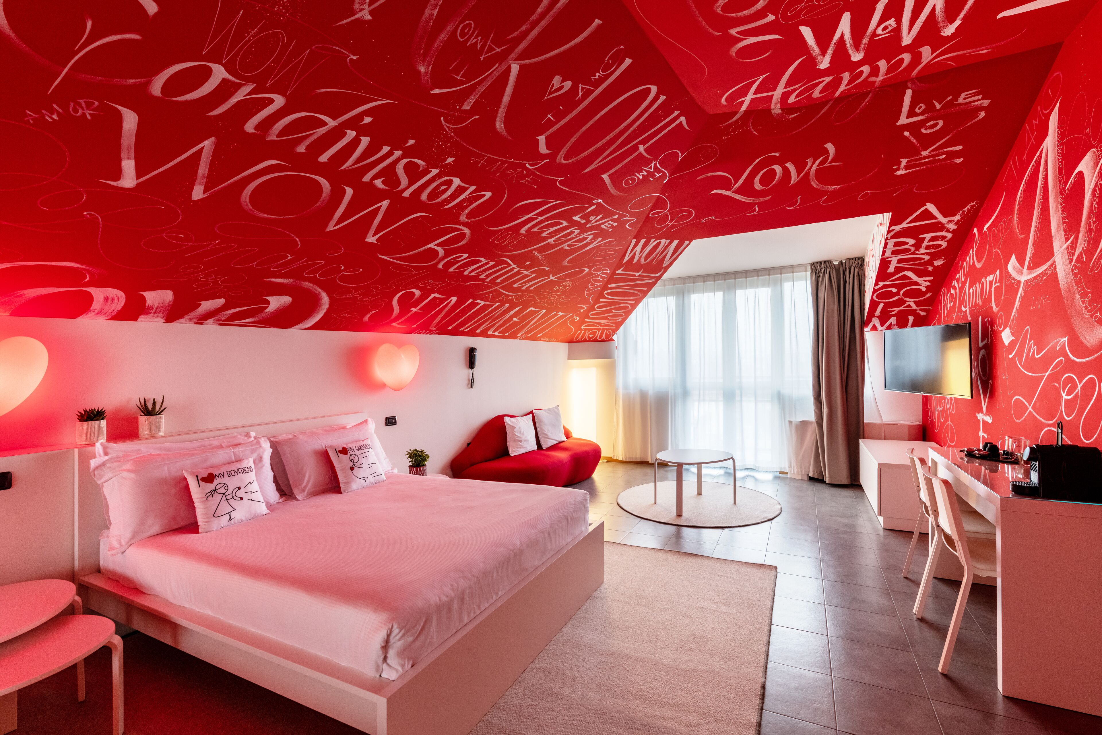 Une chambre d’hôtel moderne dotée d’un grand lit, d’un bureau et d’un coin salon. La chambre est décorée dans des tons de rouge et de blanc, et les murs sont ornés de mots inspirants.