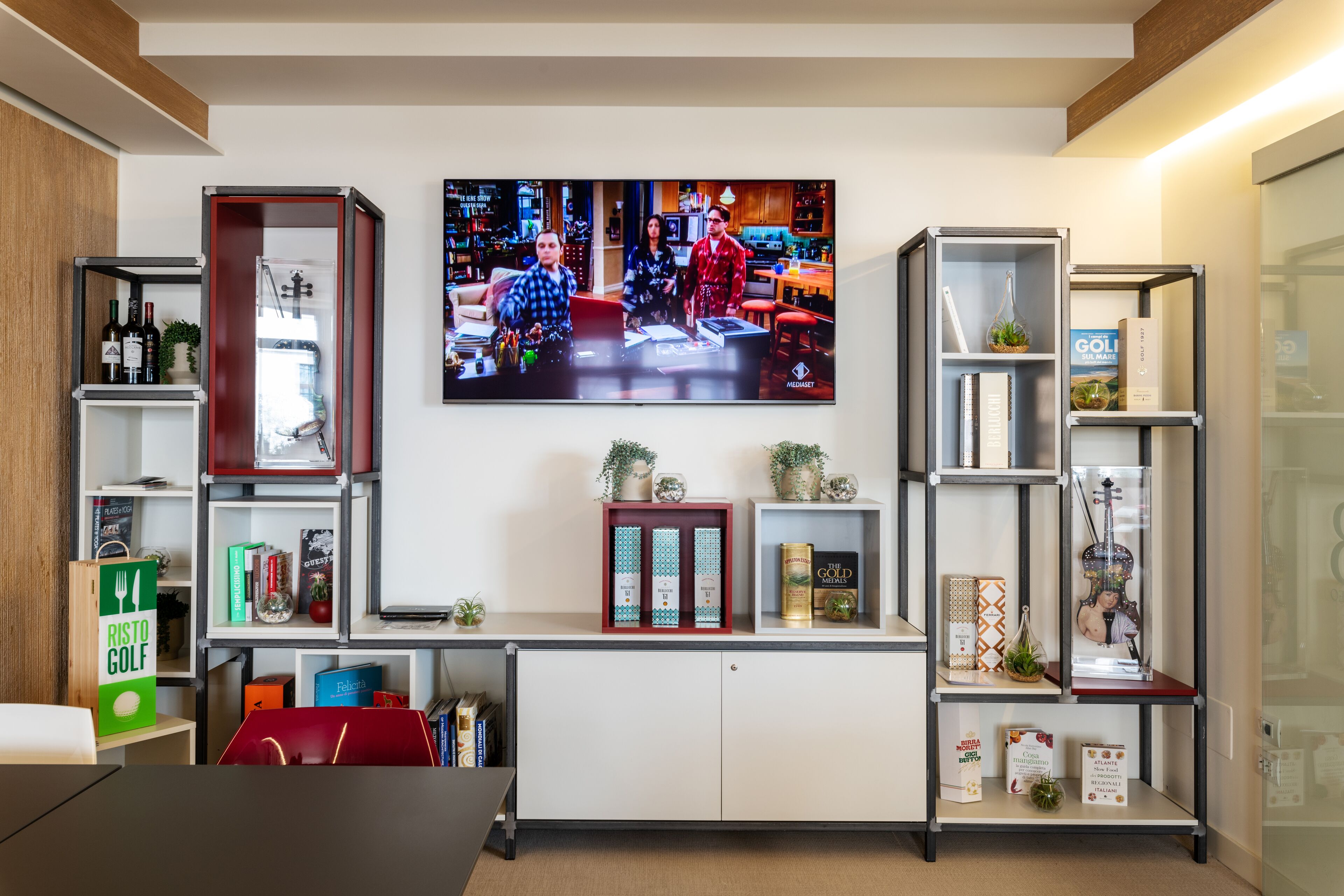 Een moderne woonkamer met een flatscreen televisie aan de muur, waarop een scène uit een programma te zien is. Er zijn boekenplanken met diverse items, waaronder boeken en decoratieve objecten. Een rode kast is in de voorgrond zichtbaar, en aan de linkerzijde is houten lambrisering te zien.