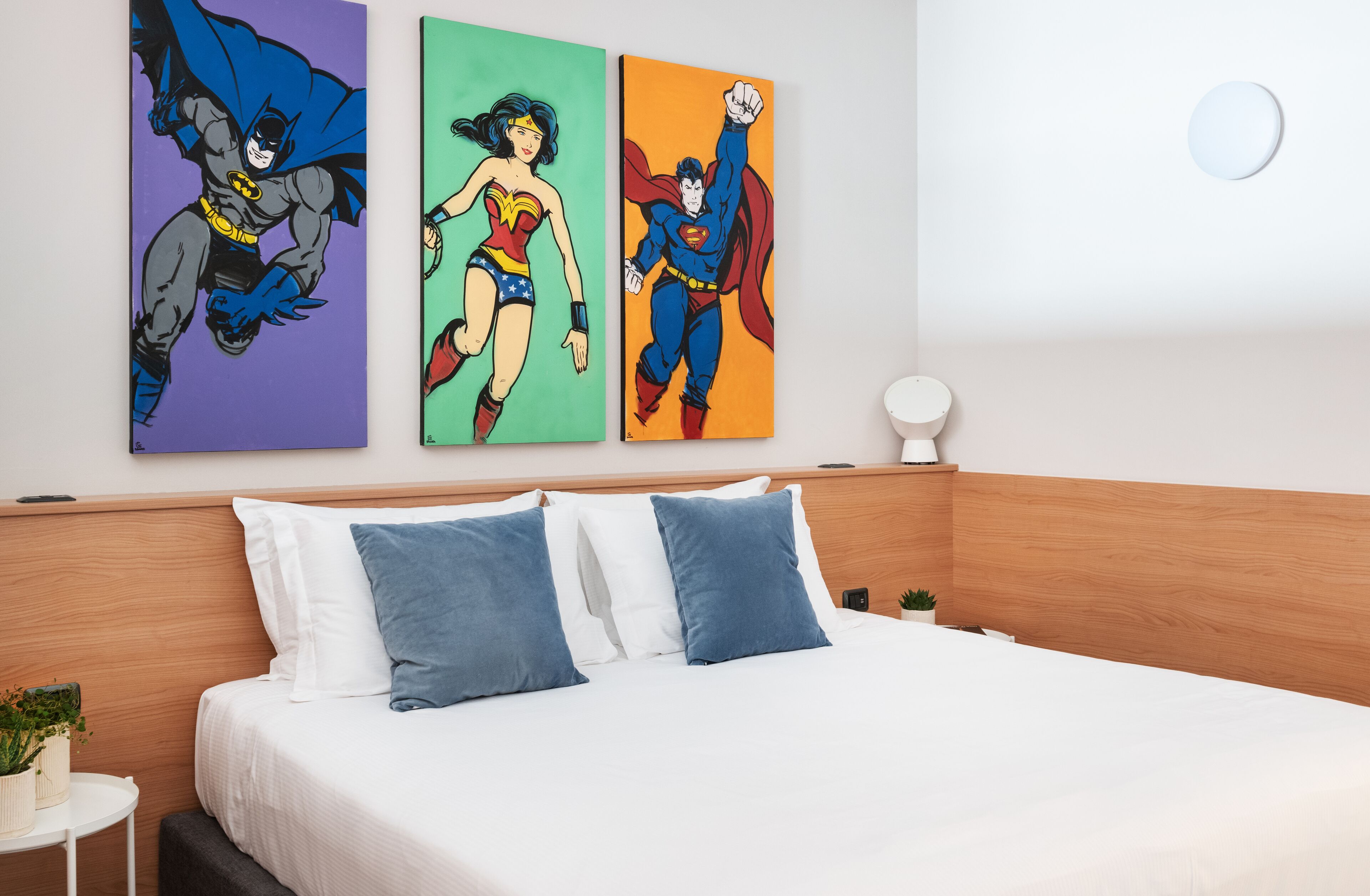 Een hotelkamer met een bed, kussens, een klein tafeltje met een plant en drie schilderijen met superheldenmotieven aan de muur.
