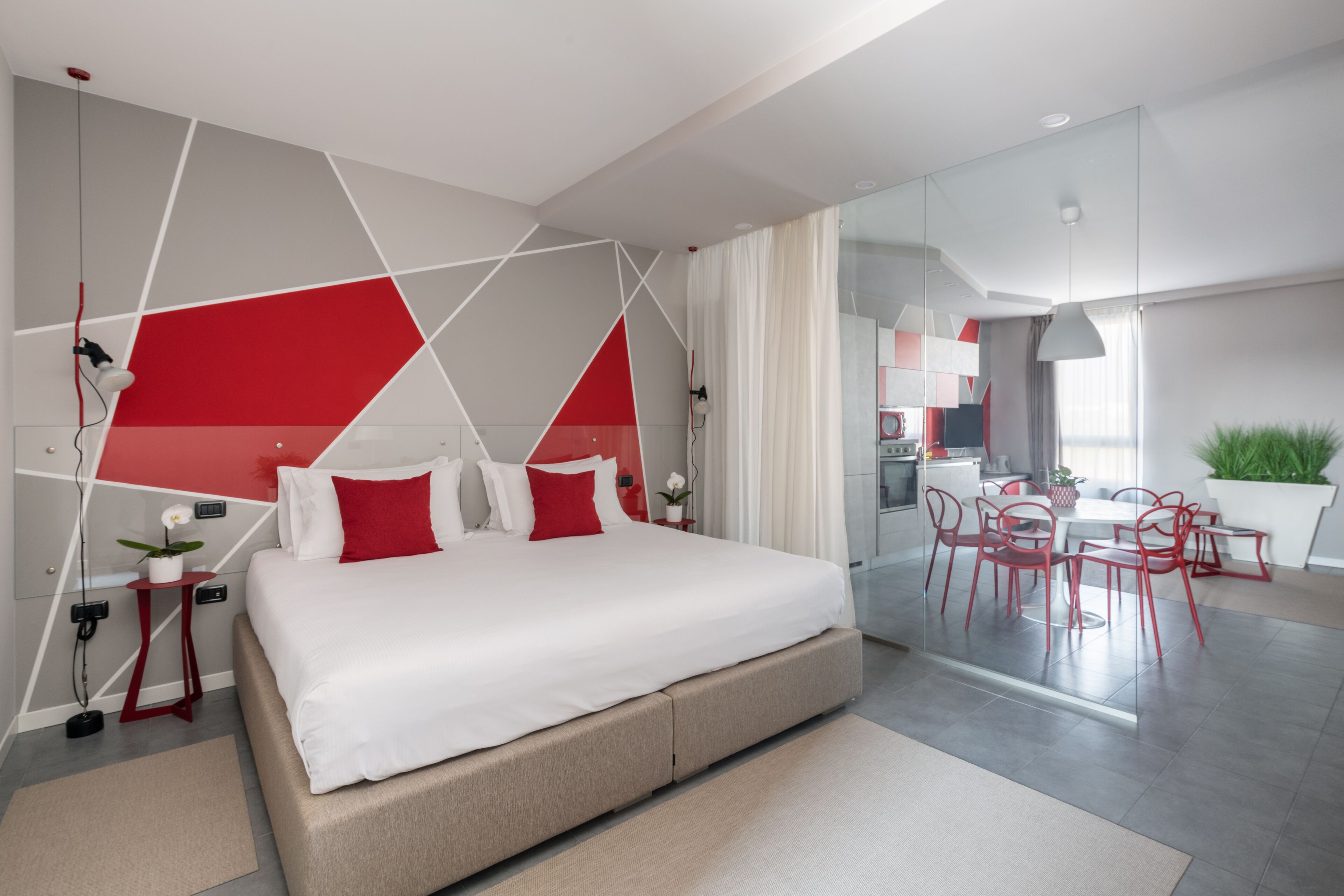 A modern bedroom with a large bed, a dining area with red chairs, and a view of the kitchen.