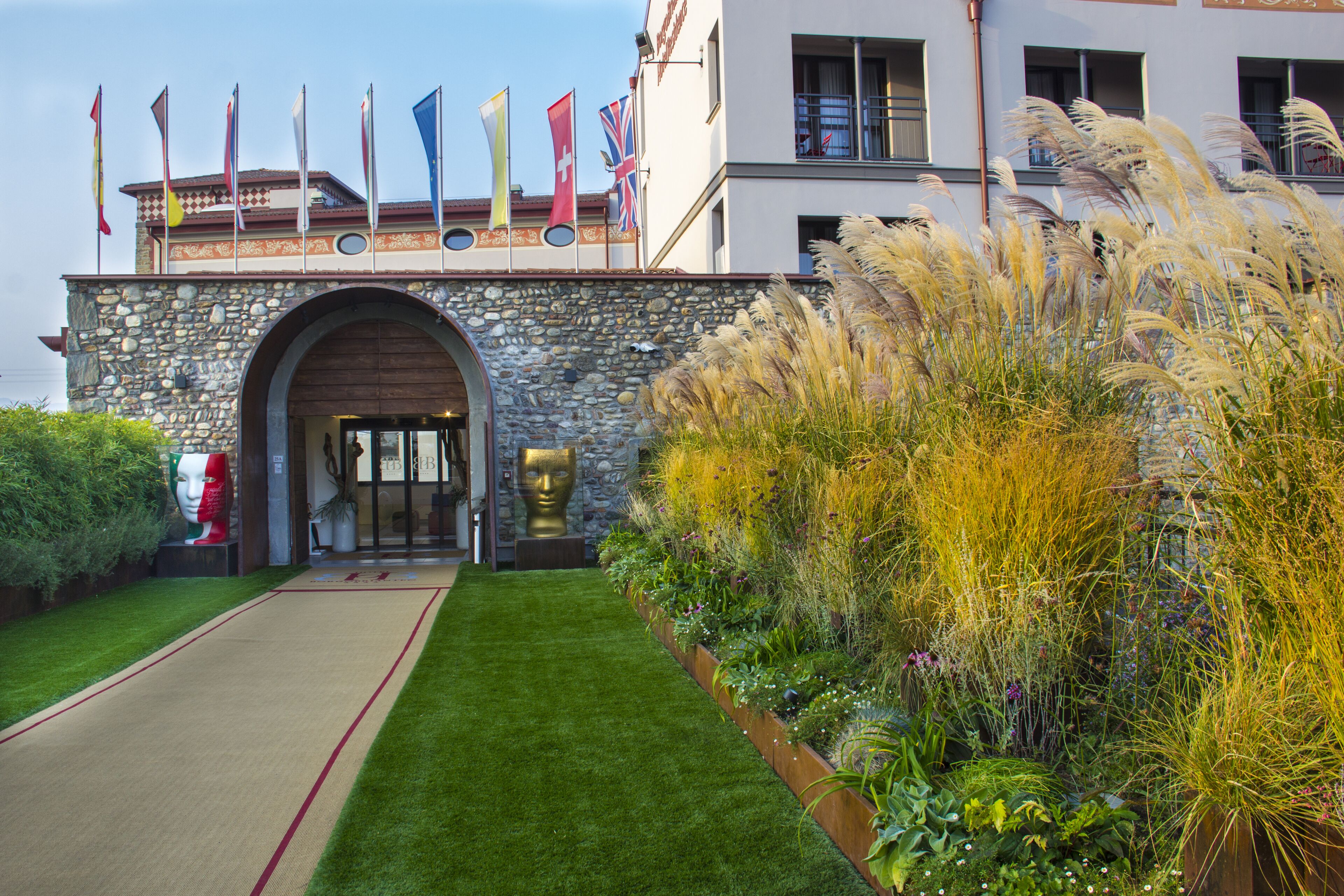Entrada de hotel con un arco de piedra, varias banderas y un jardín bien cuidado con hierbas altas.