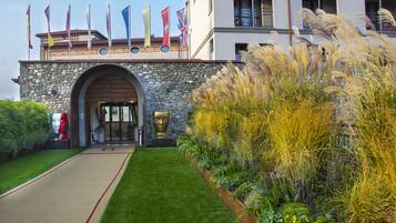 Entrada de hotel con un arco de piedra, varias banderas y un jardín bien cuidado con hierbas altas.