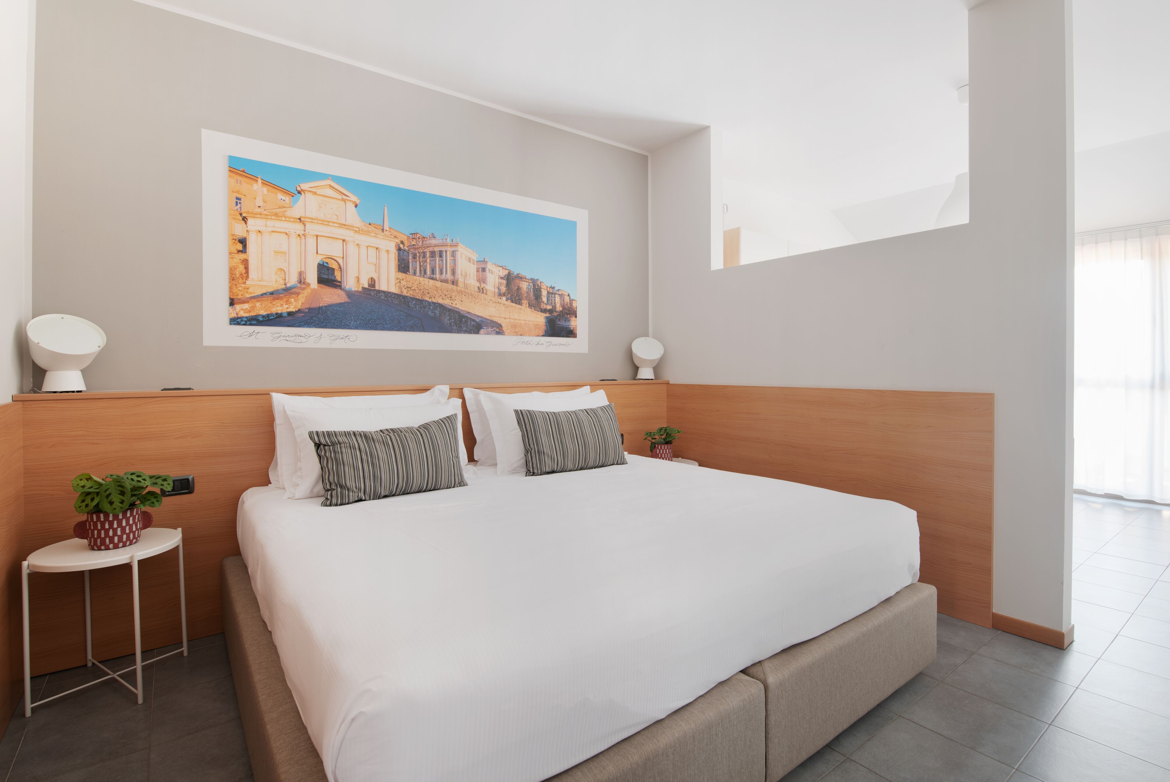 Habitación de hotel moderna con una cama grande, cabecera, mesita de noche con lámpara, planta pequeña en maceta e imagen enmarcadas en la pared.