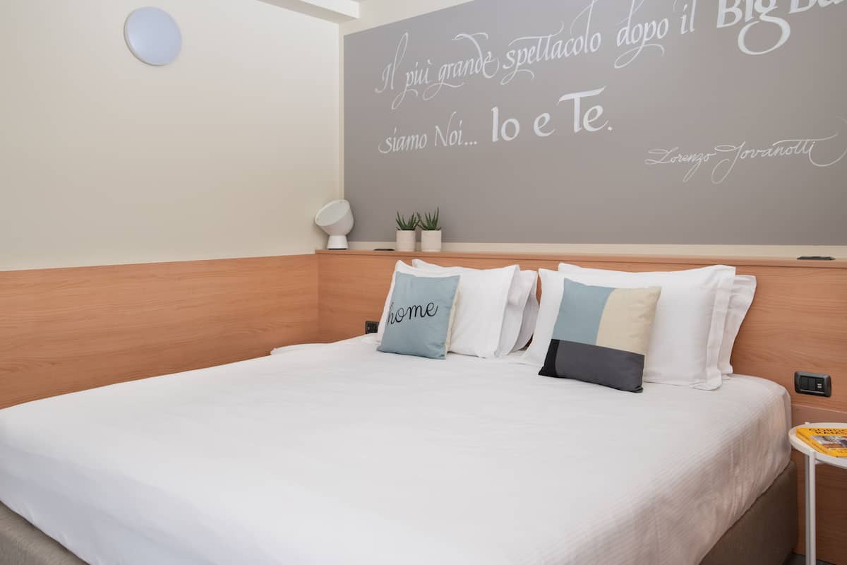 Una cama bien tendida con ropa de cama blanca y algunos almohadones, una mesita de noche con una lámpara, y una pared con una cita en italiano.