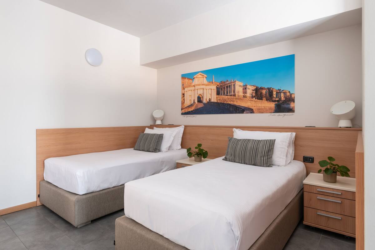 Habitación de hotel con dos camas, una mesita de noche con lámpara, un cuadro colgado en la pared y una planta en la mesita.