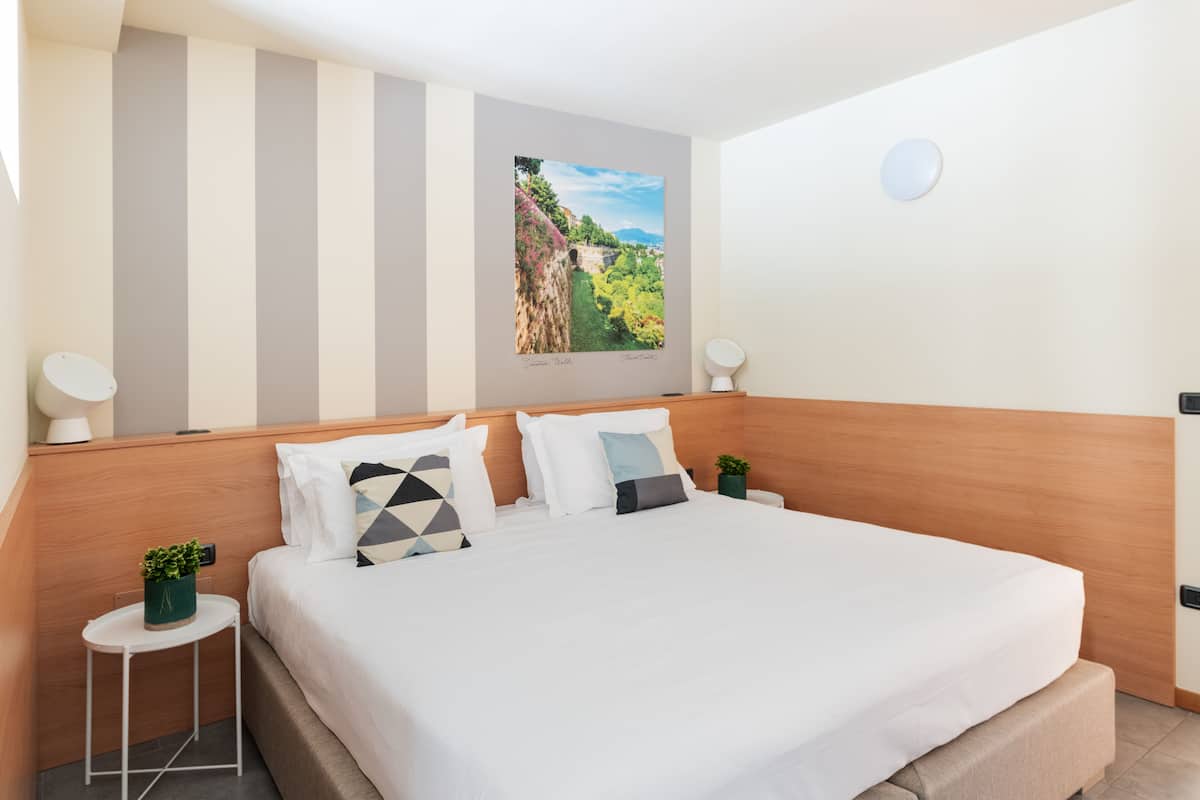 Una habitación de hotel moderna con una cama grande, paredes a rayas, un cuadro de un paisaje y una pequeña mesita de noche con una planta.