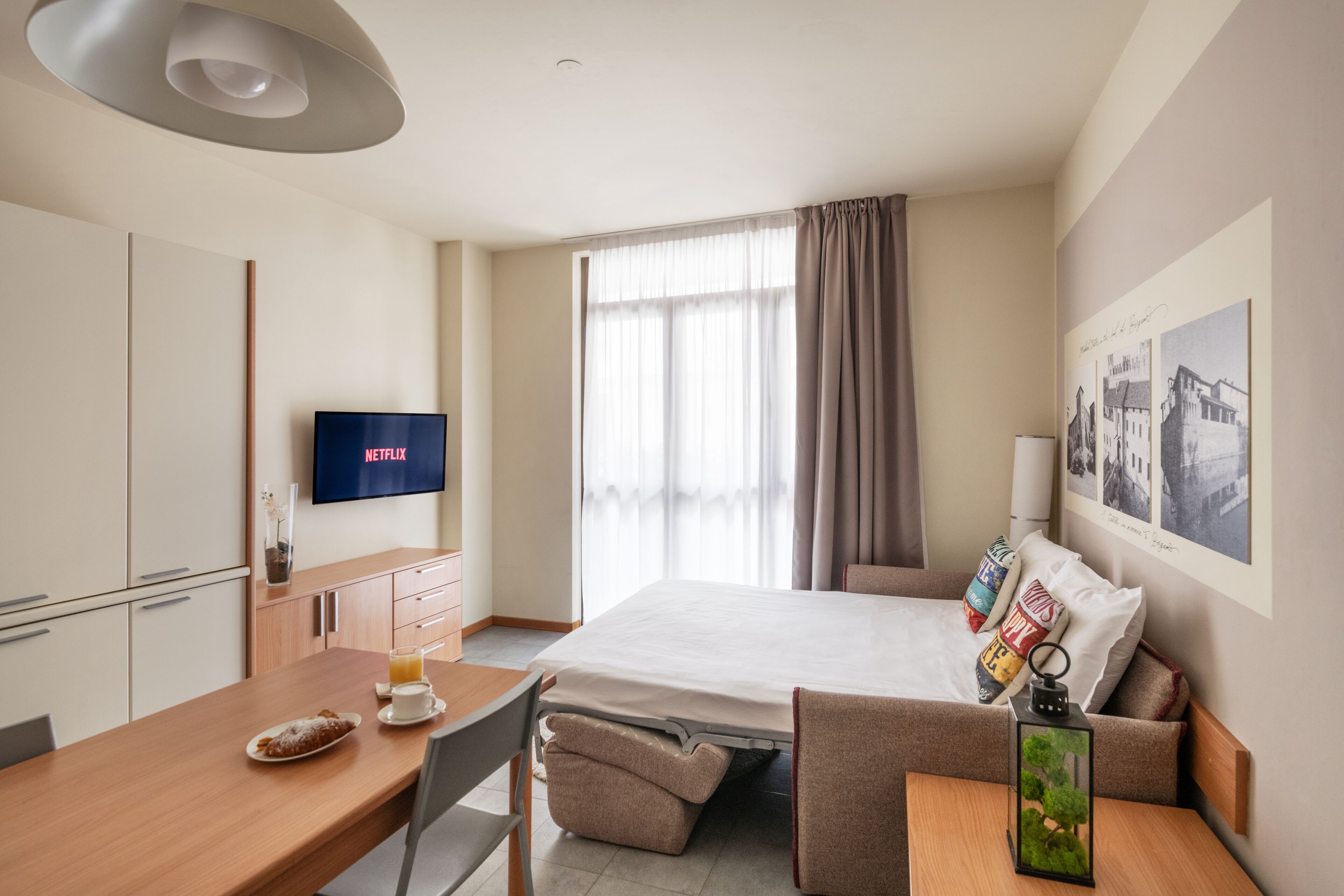A hotel room with a bed, a desk with a chair, a TV, a window with curtains, and a wall with framed pictures.
