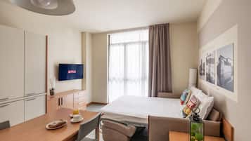 A hotel room with a bed, a desk with a chair, a TV, a window with curtains, and a wall with framed pictures.
