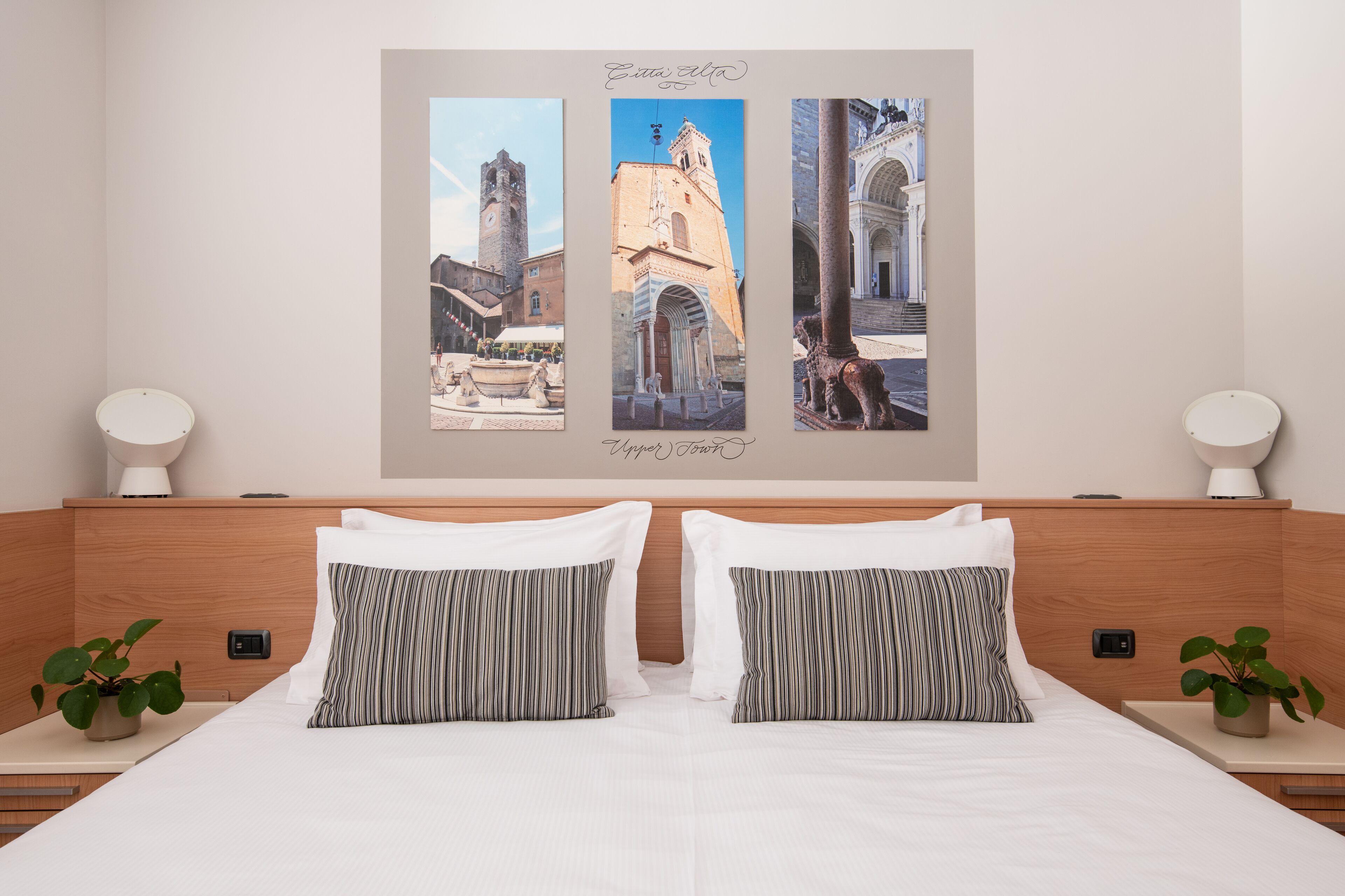 A hotel room with a large bed, two bedside tables with lamps, a wall-mounted artwork depicting a cityscape, and two potted plants.