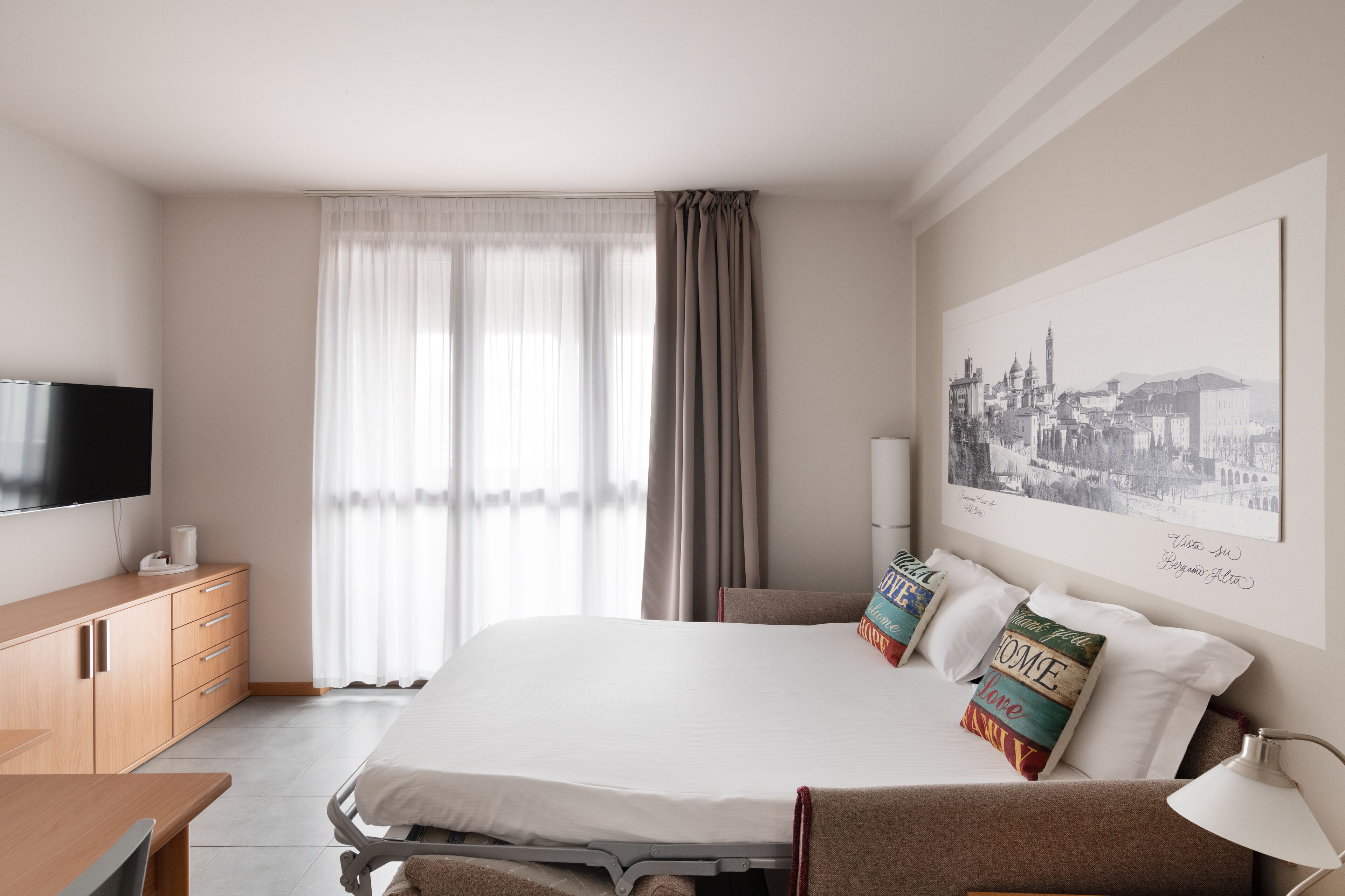 A hotel room with a bed, a TV, a desk, and a window with curtains.