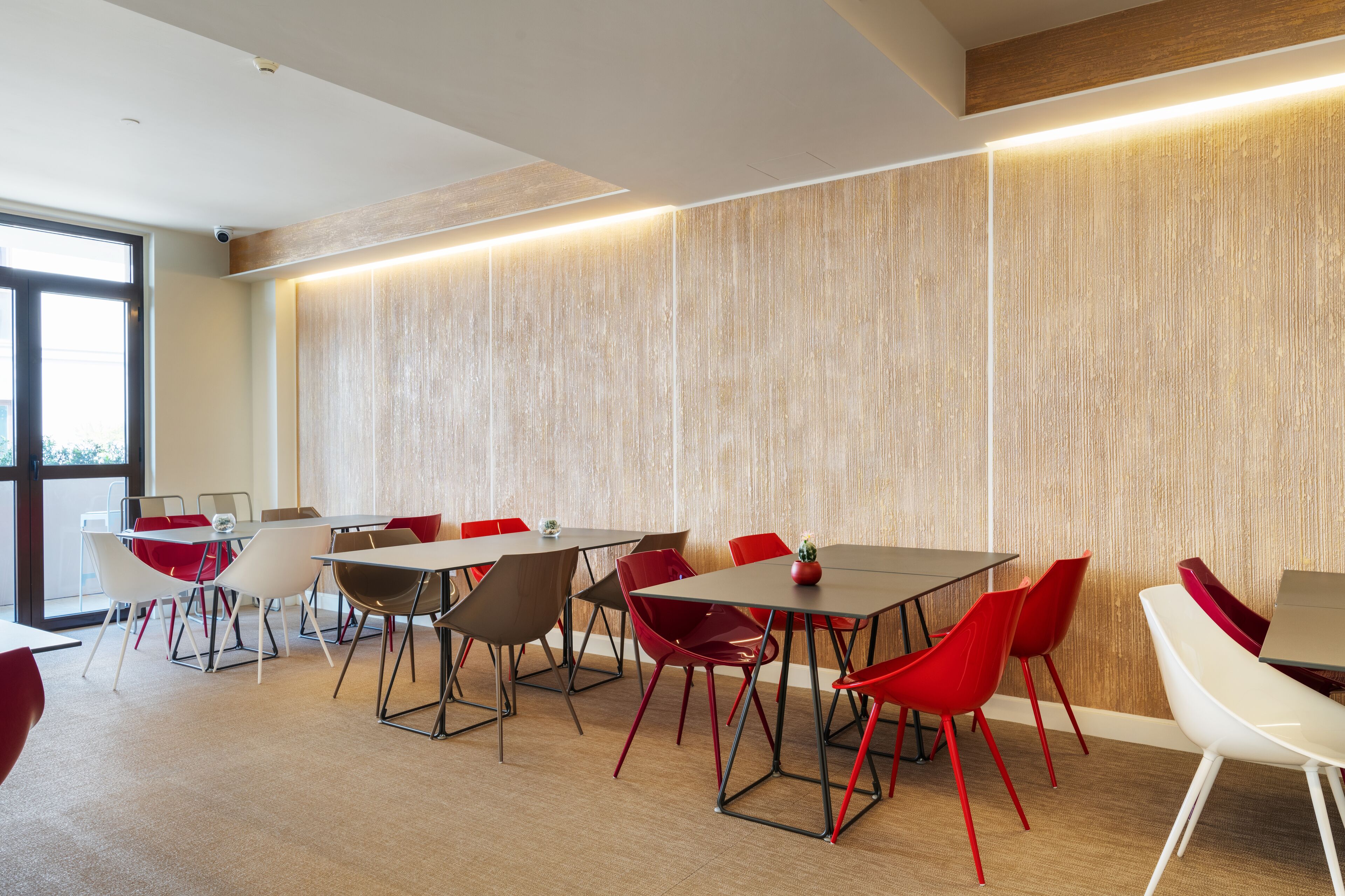 A modern conference room with multiple tables and chairs, large windows, and a wooden accent wall.