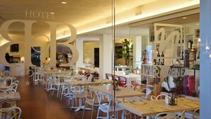 A hotel restaurant with a modern interior, featuring white chairs and tables, a glass partition, and a visible wine rack.