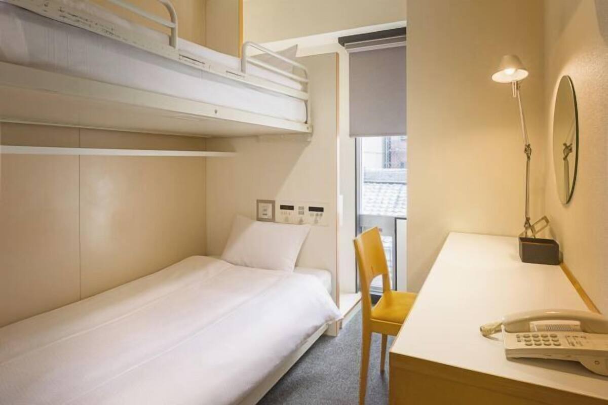 A hotel room with a bunk bed, a desk with a telephone, a chair, and a bedside table.