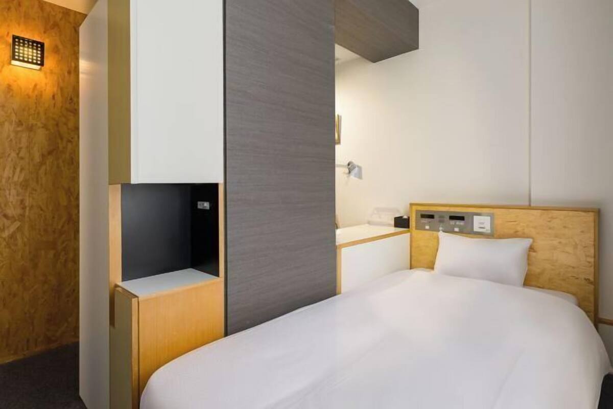 A modern hotel room with a bed, a bedside table, a control panel, and a wall-mounted light fixture.