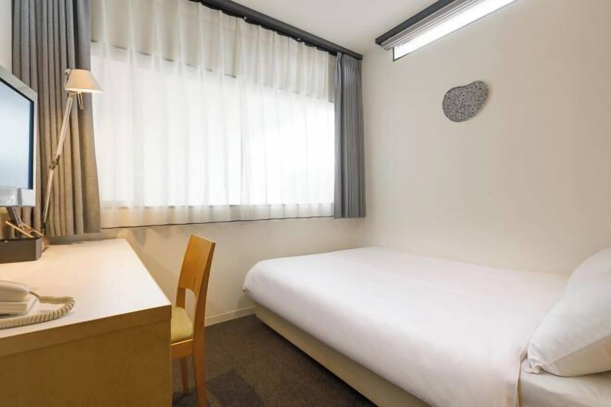A hotel room with a bed, a desk with a telephone, a chair, and a window with curtains.