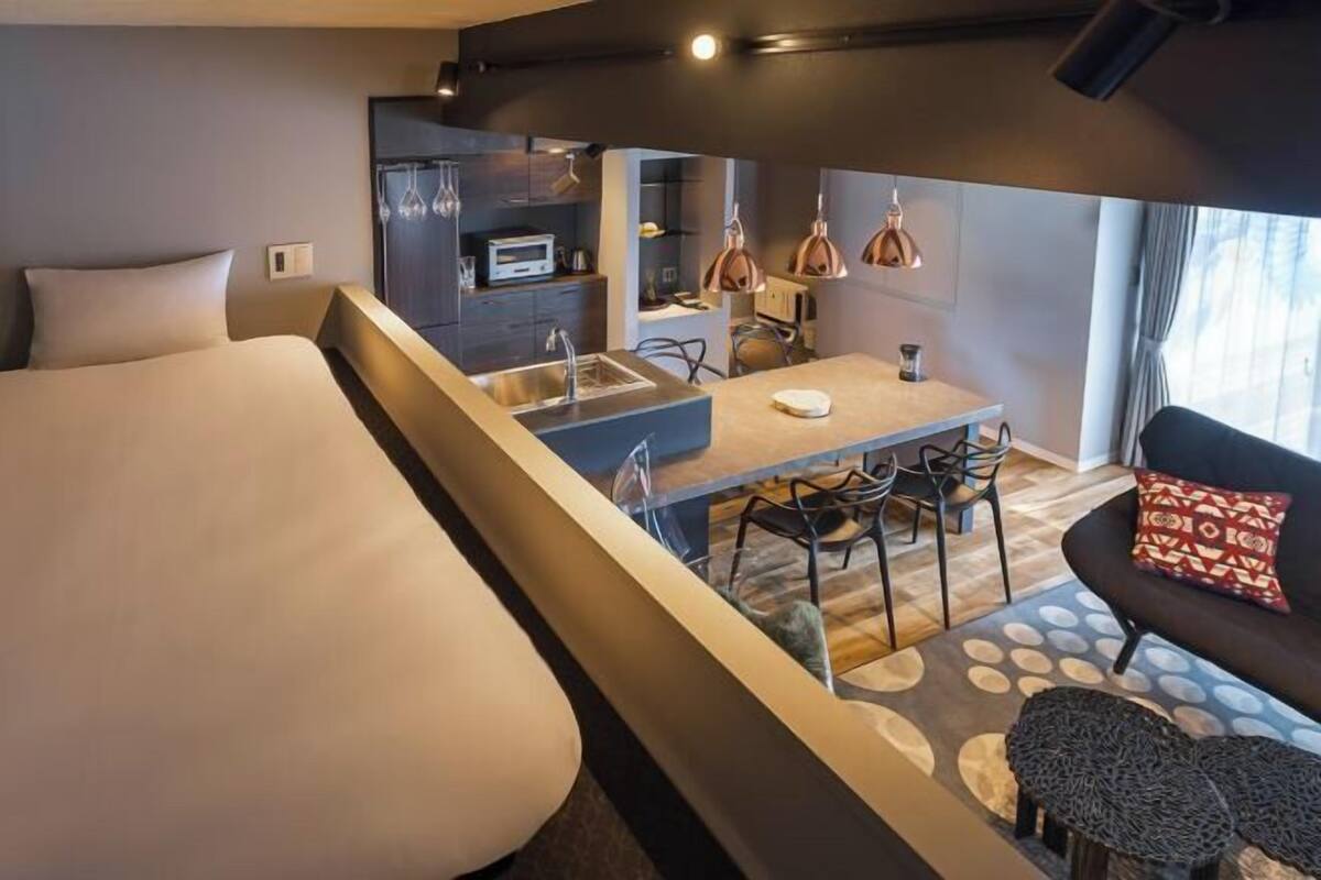 A modern hotel room with a large bed, a dining area, a kitchenette, and a seating area with a sofa and chairs.