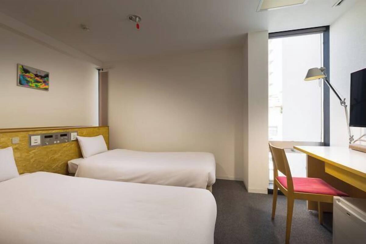 A hotel room with two beds, a desk, a chair, a TV, and a window.