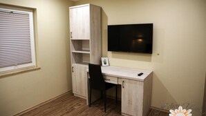 A small, well-lit room with a wooden desk, a chair, a TV mounted on the wall, and a window with blinds. - Helios Apartamenty (Ustka)