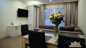 A modern living room with a sofa, a dining table with chairs, a TV mounted on the wall, and a vase of flowers. - Helios Apartamenty (Ustka)