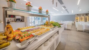 A buffet with various fruits and pastries, a digital clock, and a modern interior with white furniture and ceiling lights. - Hotel LIVVO Los Calderones - Adults Recommended (San Bartolomé de Tirajana)