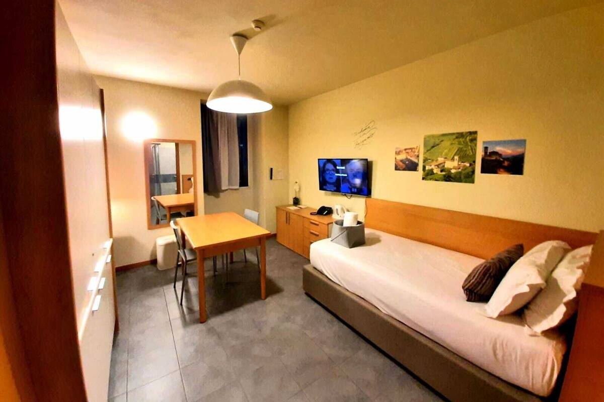 A small, well-lit room with a bed, a desk, a chair, a television, and a mirror.