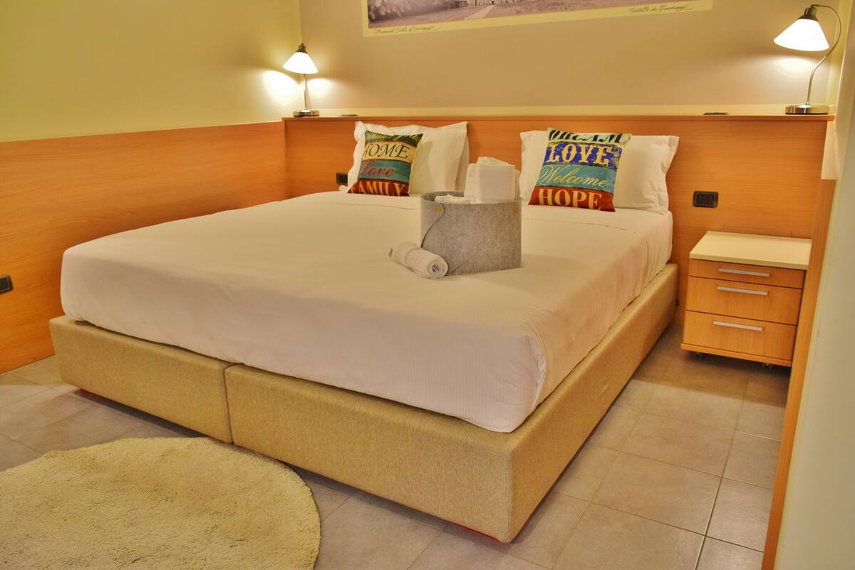 A neatly made bed with a white comforter and pillows, a bedside table with a lamp, and a small drawer unit.