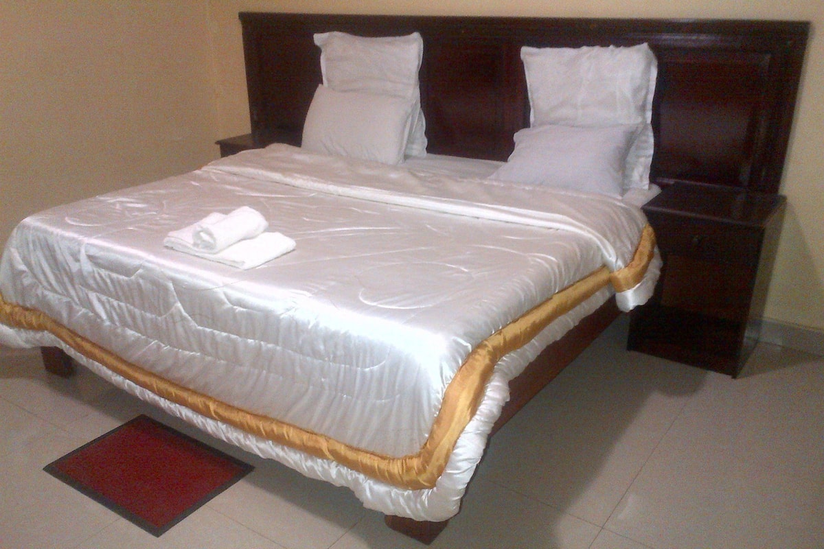 A neatly made bed with white linens and a gold trim, a wooden headboard, and a red rug on the floor.