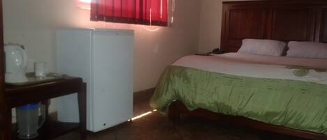 A bedroom with a bed, a small desk with a refrigerator, and a window with red curtains.