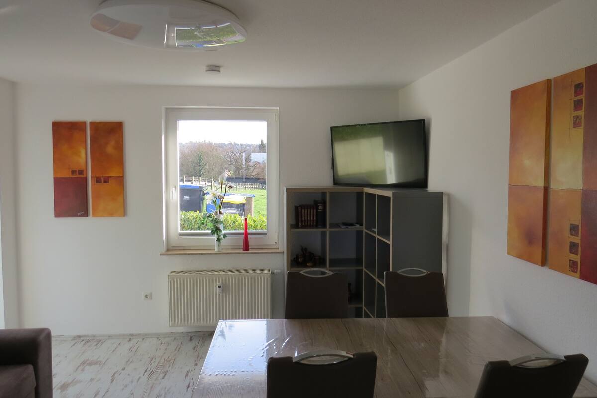 Un comedor con una mesa y sillas, un ventanal con vista a un patio, un radiador debajo de la ventana y un televisor en un estante.
