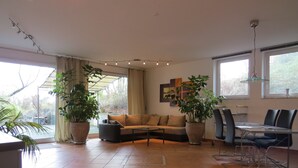 Un salón amplio con sofá, mesa de centro, mesa de comedor y sillas. Hay plantas grandes en macetas y un cuadro en la pared.