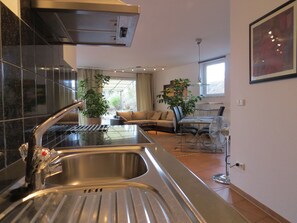 A modern kitchen with a stainless steel sink, a dining area with a table and chairs, and a living room with a sofa and a potted plant.