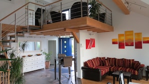 Un amplio espacio de sala de estar con techo alto, una escalera de madera, un sofá negro y una mesa de comedor.