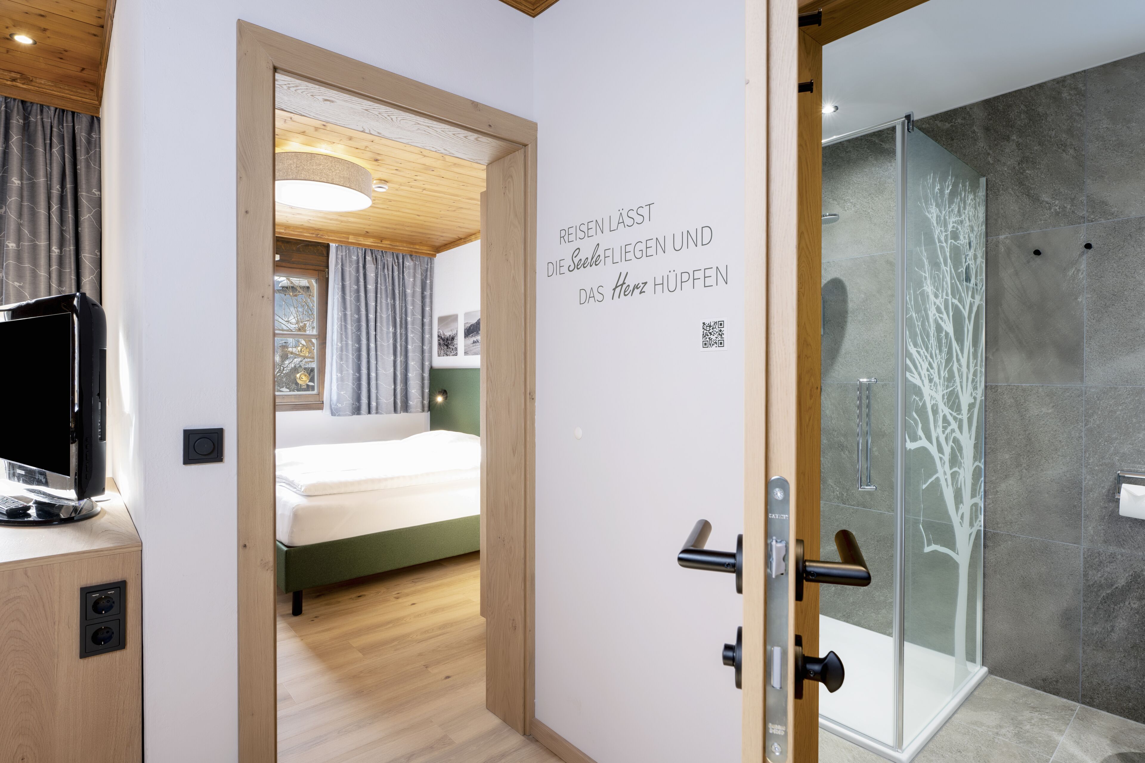 A hotel room with a bed, a television, a glass shower, and a wall with a QR code and text.