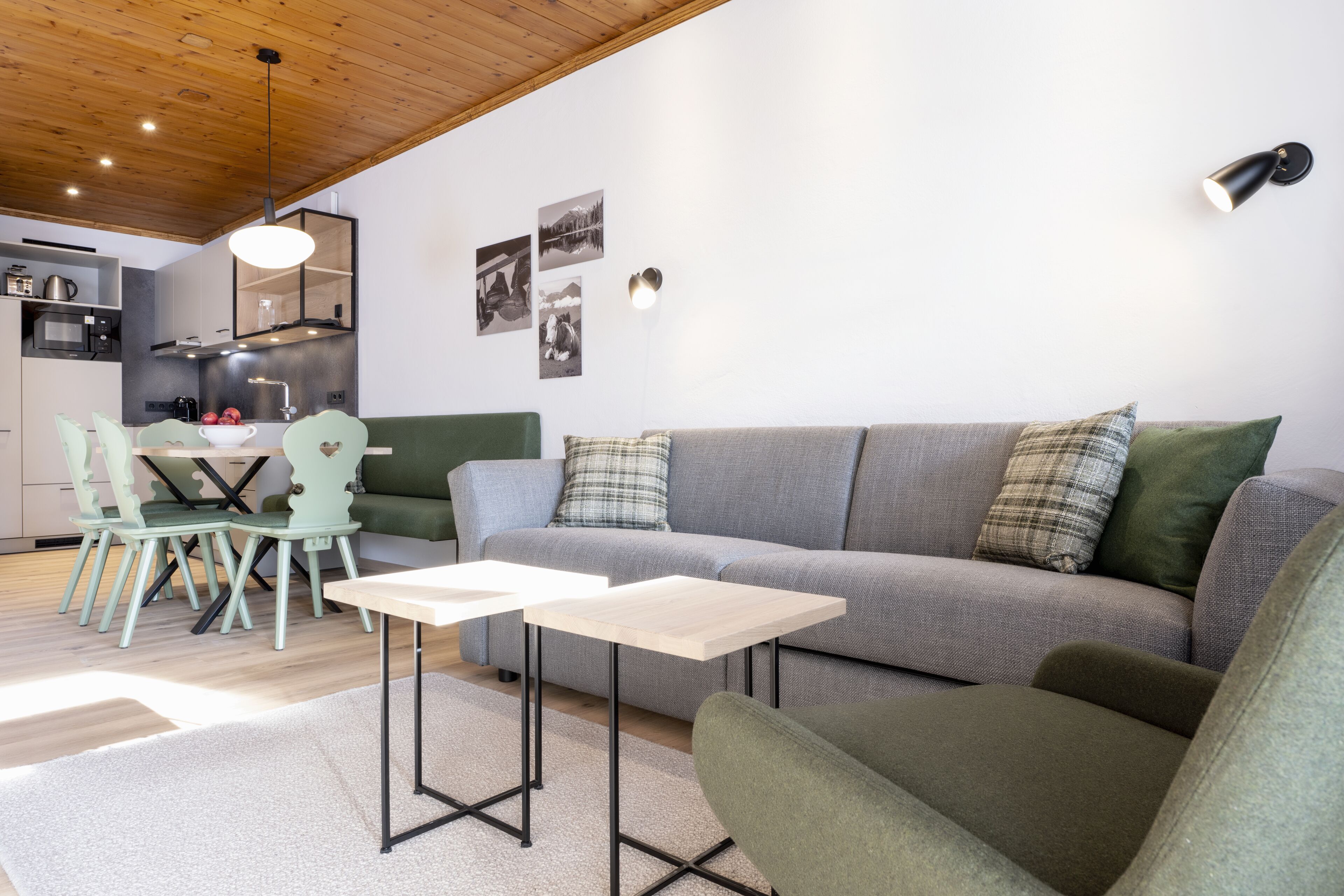 Un salon moderne avec un canapé gris, un fauteuil vert et une petite table. On y trouve également un coin repas avec une table et des chaises vertes. La cuisine est visible au fond, avec des appareils électroménagers modernes.