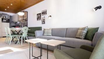 Ein modernes Wohnzimmer mit grauem Sofa, grünem Sessel und einem kleinen Tisch. Es gibt einen Essbereich mit grünem Tisch und Stühlen. Im Hintergrund ist eine Küche mit modernen Geräten zu sehen.