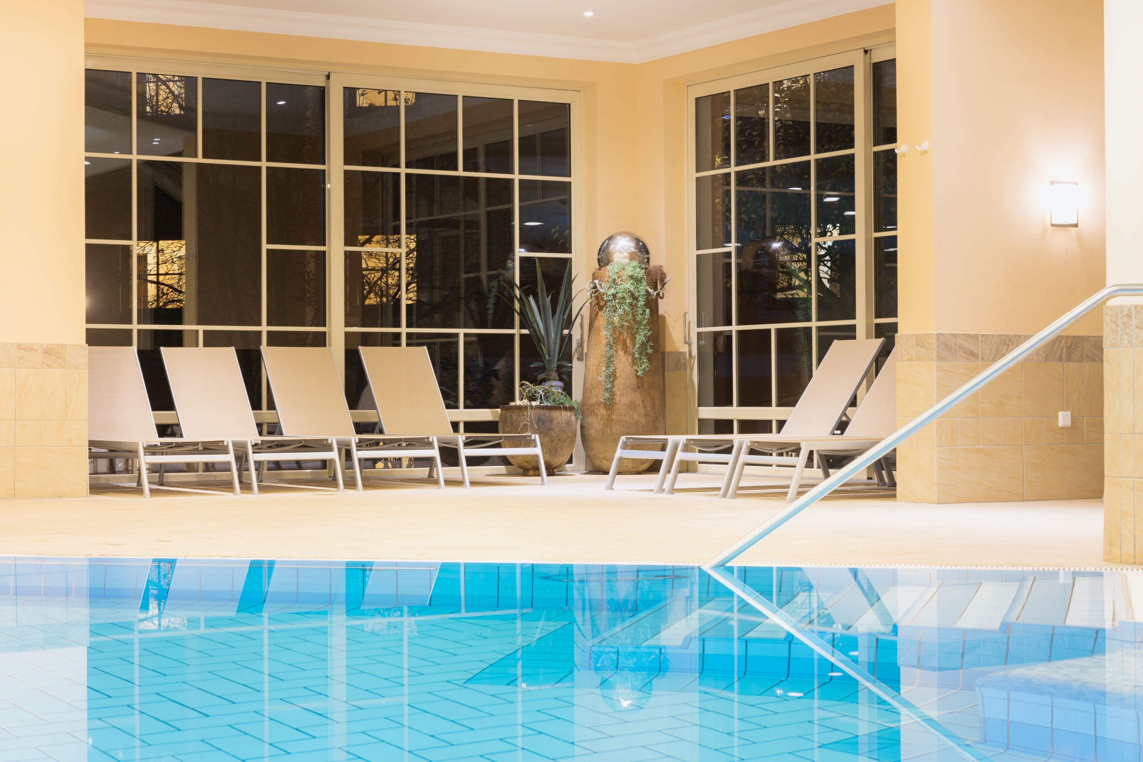 Et indendørs swimmingpool med krystalklart, blåt vand, omgivet af liggestole og store vinduer.