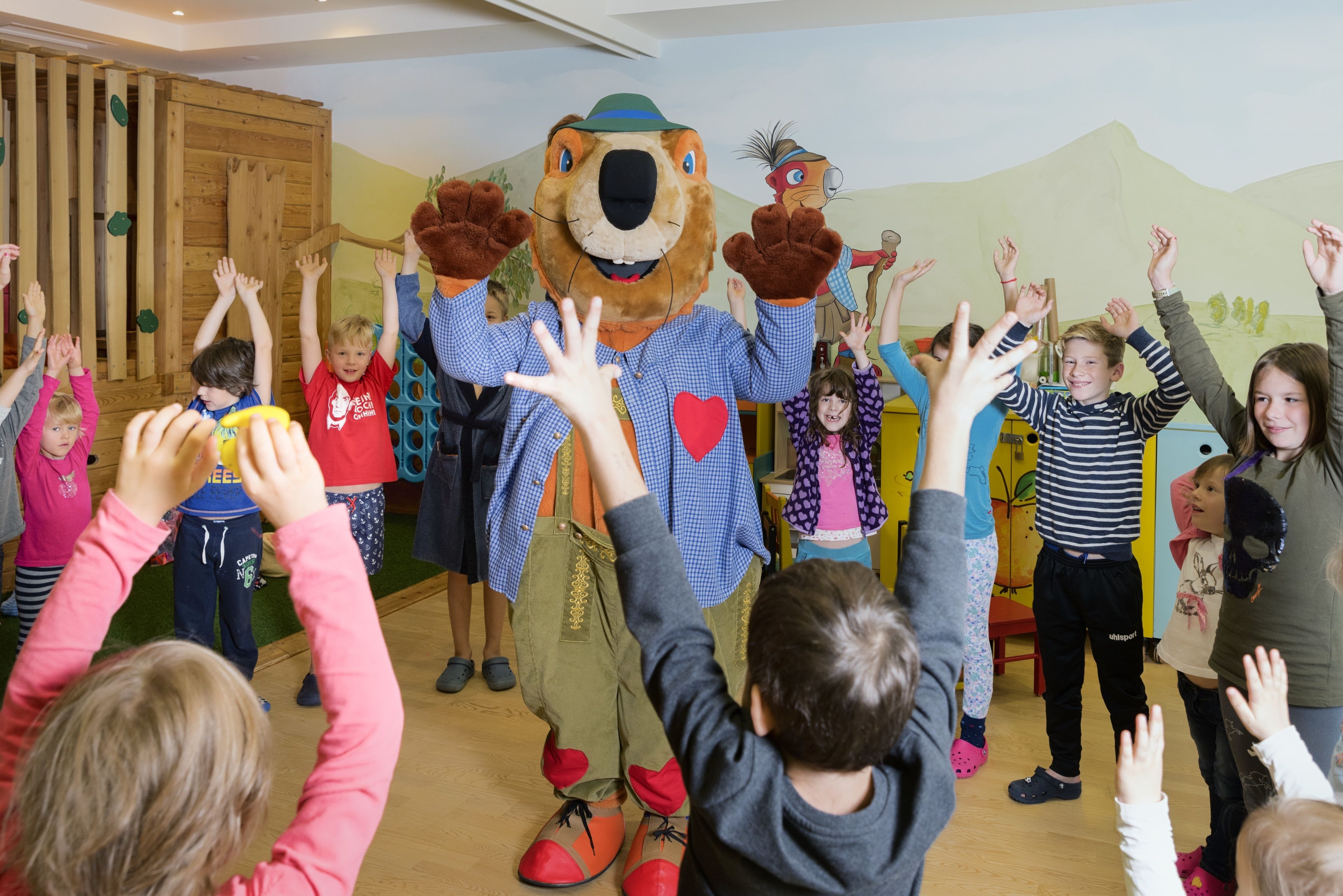 A group of children interacting with a mascot in a room with wooden flooring and a mural on the wall.
