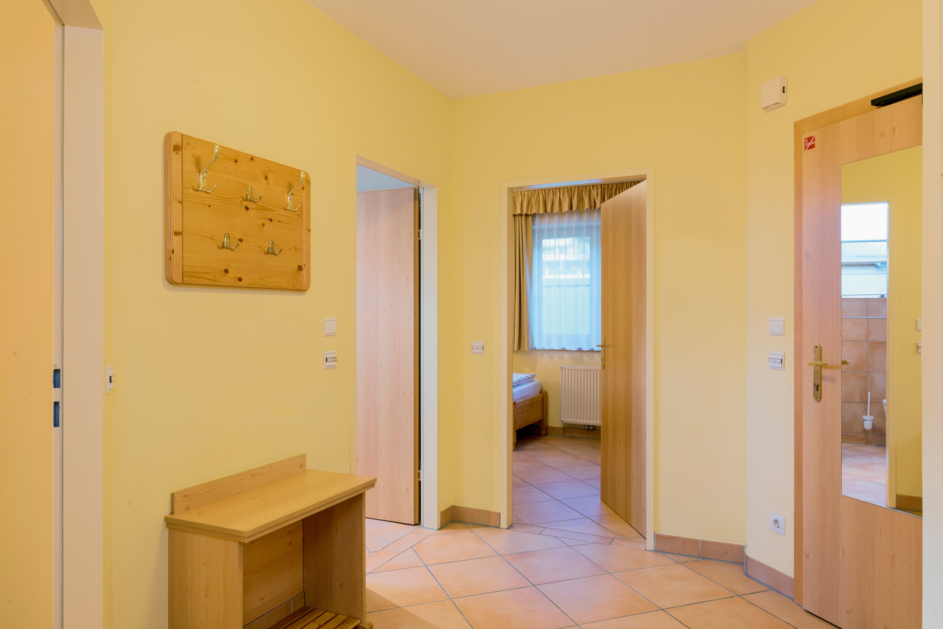 Ein Flur mit einer hölzernen Bank, einem Kleiderständer und einer Tür zu einem Schlafzimmer mit Bett und Fenster.