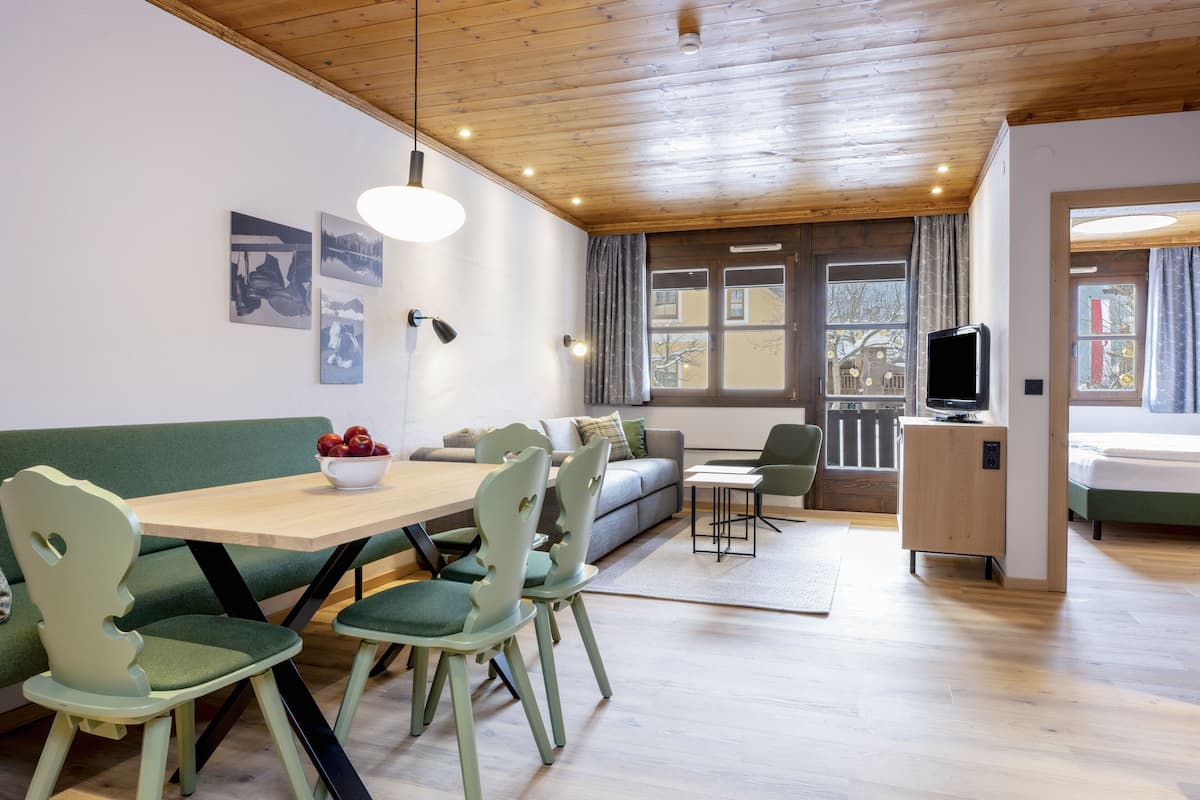 A modern living room with a wooden ceiling, a dining area with green chairs and a wooden table, a sofa, and a television.