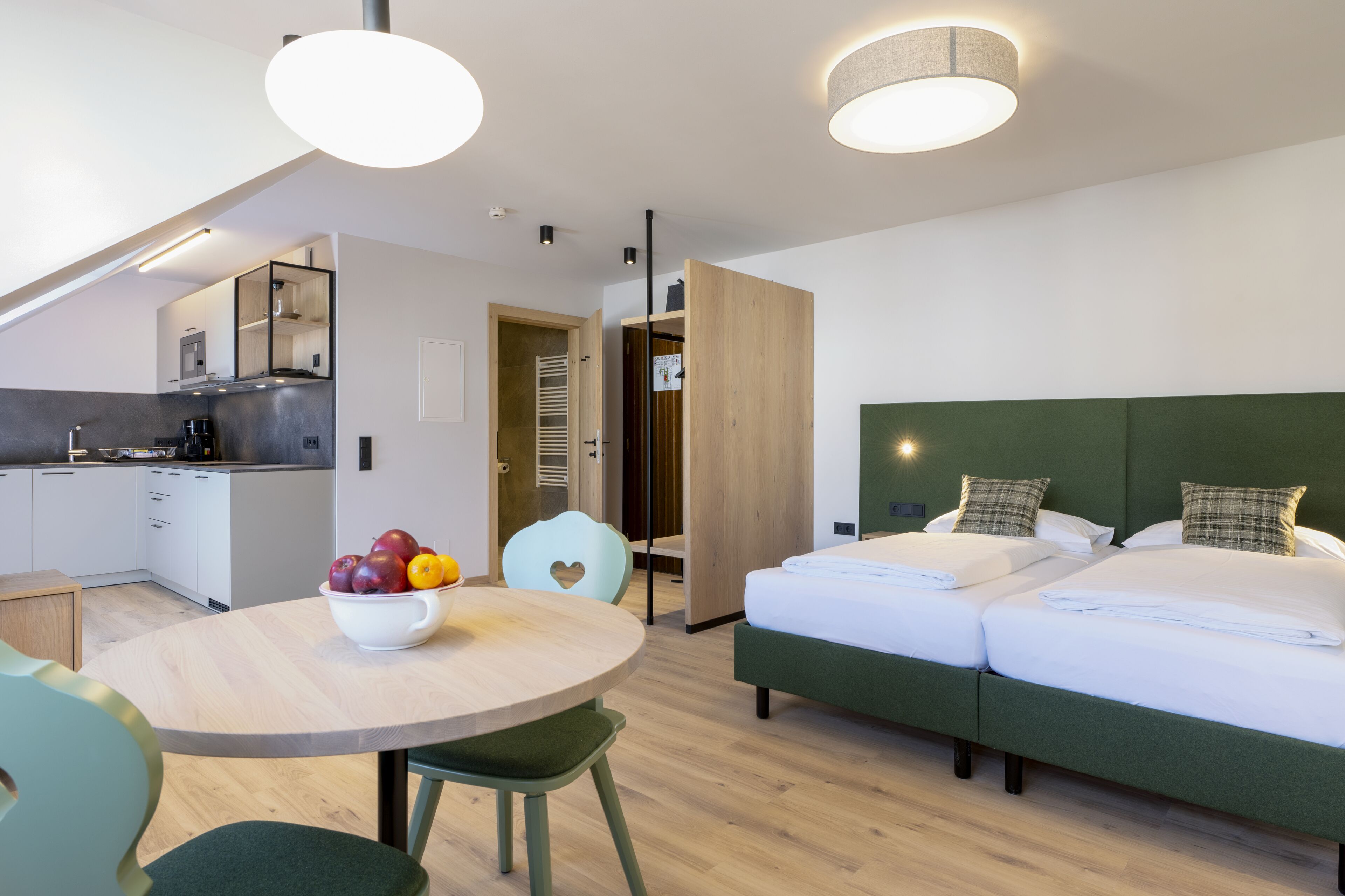 Une chambre d’hôtel moderne dotée d’une kitchenette, d’un coin repas avec un panier de fruits, d’un lit avec une tête de lit verte et d’une vue sur la ville.