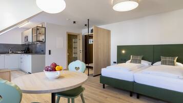 Une chambre d’hôtel moderne dotée d’une kitchenette, d’un coin repas avec un panier de fruits, d’un lit avec une tête de lit verte et d’une vue sur la ville.